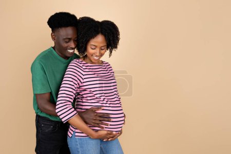 Téléchargez les photos : Joyeux jeune homme noir étreignant dame avec gros ventre, sentir le mouvement du bébé, profiter de la grossesse et attendre enfant, isolé sur un fond beige, studio. Parents et nouvelle vie, famille - en image libre de droit