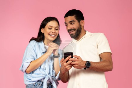 Téléchargez les photos : Magnifique jeune couple hispanique utilisant un smartphone sur fond rose, beau mec millénaire montrant sa jolie petite amie excitée belle offre en ligne, les amateurs de shopping sur Internet ensemble - en image libre de droit