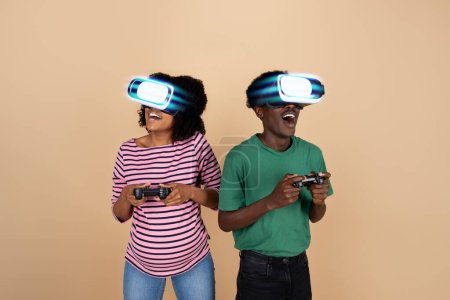 Téléchargez les photos : Heureux surpris jeune famille noire avec ventre, en lunettes VR jouer jeu en ligne avec des joysticks, isolé sur fond beige, studio, collage. Amusement et divertissement dans le temps libre, émotions avec l'appareil - en image libre de droit