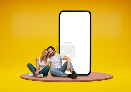 Photo for Happy cheerful beautiful young couple sitting on platform over yellow studio background, embracing next to huge phone with white blank screen, mockup for onlne offer summer deal, copy space - Royalty Free Image