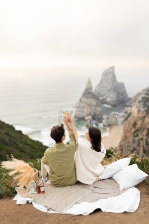 Photo for Back view of romantic couple celebrating their engagement or honeymoon on the coastline, drinking champagne and clinking glasses, enjoying ocean shore, copy space, vertical shot - Royalty Free Image