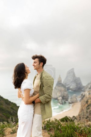 Téléchargez les photos : Jeune couple amoureux ayant rendez-vous romantique, s'embrassant et se regardant, debout sur le littoral avec une belle vue sur la côte de l'océan, plan vertical, espace de copie - en image libre de droit