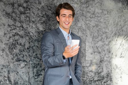 Téléchargez les photos : Portrait de joyeux beau jeune homme d'affaires portant costume en utilisant le téléphone et le sourire, debout près du mur de marbre gris, en utilisant la plus récente application mobile d'affaires ou chatbot assistant en ligne, espace de copie - en image libre de droit