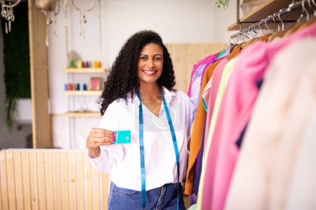 Photo for Successful fashion business. Cheerful middle eastern clothing designer lady showing credit card posing with handcrafted garments on rack at dressmaking atelier interior. Commerce and profit concept - Royalty Free Image