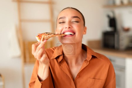 Foto de Mujer caucásica joven contenta disfrutando de su creación de cocina, mordiendo rebanada de pizza casera, cenando, sentado en la mesa en la cocina, espacio libre, primer plano - Imagen libre de derechos