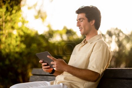 Téléchargez les photos : Jeune homme nomade numérique assis sur le banc au parc public, en utilisant une tablette numérique, lecture blog sur e-book, espace de copie. Happy millennial guy websurfing, défilement, en utilisant une belle application mobile, se reposant en plein air - en image libre de droit