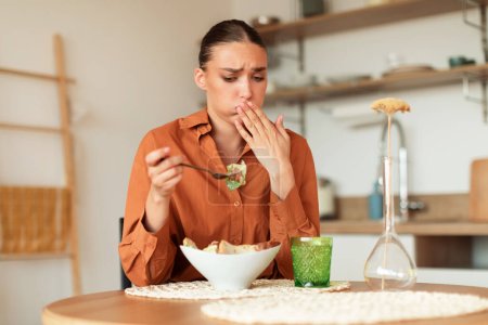 Photo for Young caucasian woman feeling nausea while eating delivered caesar salad, lady looking at plate and closing mouth, sitting at table in kitchen interior - Royalty Free Image