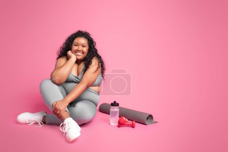 Photo for Recovery after workout training. Happy black body positive woman resting on floor with water bottle, dumbbells and mat on pink background, free space - Royalty Free Image