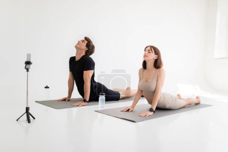Photo for Calm young caucasian lady, guy enjoy workout together, meditate, practice yoga, body stretching, shoot video on phone in gym or room interior. App for sports blog at home, health care with coach - Royalty Free Image