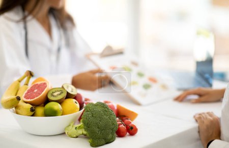 Mature caucasian doctor nutritionist in white coat recommends to young woman diet plan at table with organic vegetables and fruits in office interior. Weight loss, professional advice