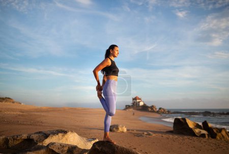 Foto de Mujer caucásica milenaria positiva practica yoga, disfruta de ejercicios de respiración, estiramiento de piernas en la playa del mar. Entrenamiento matutino, deportes, cuidado de la salud y fitness solo, larga duración - Imagen libre de derechos