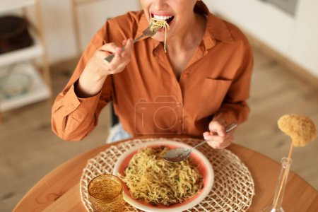 Téléchargez les photos : Jeune femme caucasienne mangeant de savoureuses pâtes italiennes au parmesan, savourant un délicieux repas assis à table à l'intérieur de la cuisine, rogné, gros plan - en image libre de droit