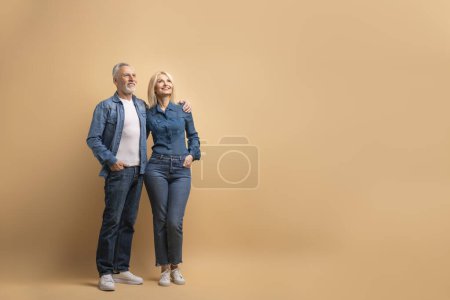 Photo for Loving cheerful beautiful senior couple wearing casual denim outfit embracing and looking at copy space, isolated on beige background. Elderly man and woman checking nice advertisement - Royalty Free Image