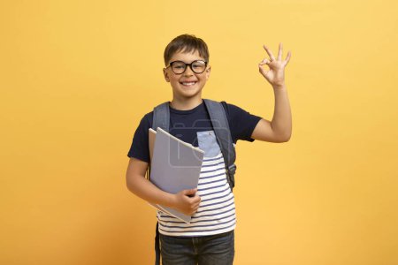Photo for Happy cheerful caucasian boy schooler with backpack behind his back and books in his hand showing okay gesture and smiling, isolated on yellow background, copy space. Great offer for schoolers - Royalty Free Image