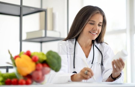 Photo for Positive european mature lady doctor nutritionist makes notes, diet program, look at jar of pills, vitamins, food supplements at table with fruits in clinic interior. Work professional, health care - Royalty Free Image