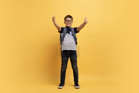 Photo for Happy cheerful caucasian kid boy schooler with backpack behind his back showing thumb ups and smiling, isolated on yellow background, copy space, full length. Great offer for schoolers - Royalty Free Image
