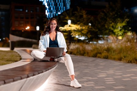 Téléchargez les photos : Femme pigiste travaillant sur un ordinateur portable moderne, assise sur un banc la nuit, travaillant avec un ordinateur dans un parc urbain sur fond urbain moderne, espace libre - en image libre de droit