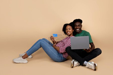Photo for Cheerful young black man and lady with big belly sit on floor, enjoy pregnancy with laptop, show credit card, isolated on beige background, studio. Parents and family and online shopping, banking - Royalty Free Image