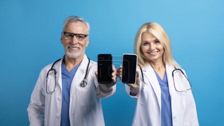Friendly cheerful senior doctors man and woman in medical white coats showing smartphones with black empty screen, great mobile app for telemedicine, blue background, mockup