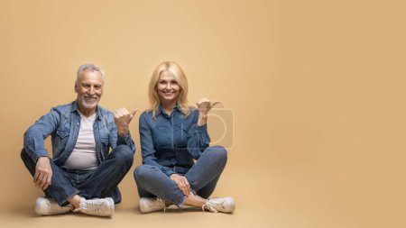Photo for Positive cheerful beauitufl elderly man and woman in casual denim outfit sitting on floor isolated on beige background, pointing at copy space. Happily married senior couple showing advertisement - Royalty Free Image