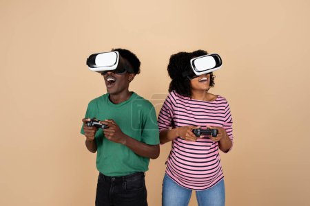 Téléchargez les photos : Joyeux surpris jeune homme noir et dame avec gros ventre, lunettes VR jouer jeu en ligne avec des joysticks, isolé sur fond beige, studio. Relations, amusement et divertissement dans le temps libre - en image libre de droit