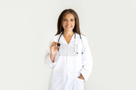 Téléchargez les photos : Smiling européenne femme d'âge moyen médecin nutritionniste en manteau blanc tient stéthoscope, écoute la respiration, isolé sur fond studio lumière. Travail professionnel et soins de santé, traitement et examen - en image libre de droit