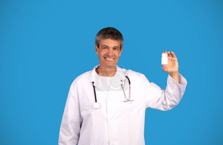 Téléchargez les photos : Homme médecin souriant montrant bouteille médicale avec des pilules médicaments publicitaires sur fond bleu, en regardant la caméra. Médecin généraliste avec stéthoscope recommandant un traitement médical - en image libre de droit