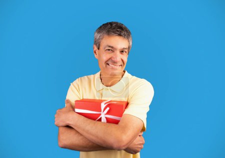 Photo for Holiday surprise. Funny mature man holding and hugging present box, celebrating his birthday and receiving holiday gifts posing on blue studio background, smiling at camera - Royalty Free Image