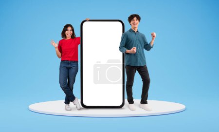 Photo for Excited young middle eastern woman and european woman standing by big phone with white blank sceen, blank space for advertisement, posing over blue background, couple recommending great online offer - Royalty Free Image