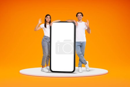 Photo for Positive smiling stylish millennial man and woman friends couple standing on platform with huge phone with white blank screen and showing okay, orange background. Great mobile app, mockup - Royalty Free Image