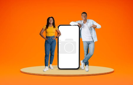 Foto de Linda joven pareja afroamericana alegre de pie en la plataforma sobre fondo naranja, mostrando una buena oferta en línea en un gran teléfono inteligente con pantalla vacía blanca, espacio en blanco, maqueta - Imagen libre de derechos