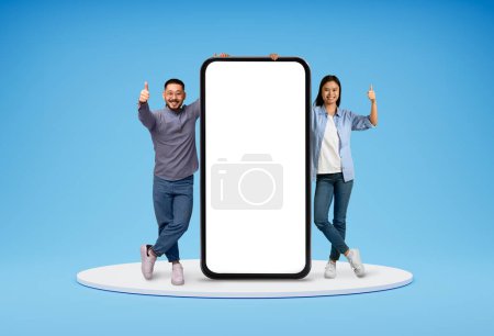 Photo for Positive smiling stylish millennial asian man and woman friends couple standing on platform with huge phone with white blank screen and showing thumb up, blue background. Great mobile app, mockup - Royalty Free Image