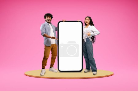 Foto de Lindo feliz sonriente joven indio pareja hombre y mujer de pie en la plataforma redonda por teléfono grande con pantalla blanca en blanco, fondo de estudio rosa, espacio en blanco maqueta para aplicación móvil, collage - Imagen libre de derechos