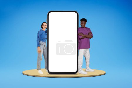 Photo for Beautiful couple cheerful young woman and black guy boyfriend showing great online deal, standing on platform by phone with white blank screen, blue studio background, empty space, mockup - Royalty Free Image