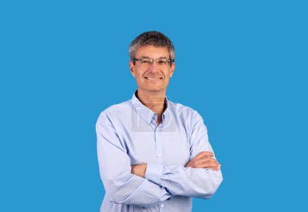 Photo for Confident business person. Happy mature businessman in shirt posing and crossing hands with expression of confidence, smiling looking at camera standing over blue background, studio shot - Royalty Free Image