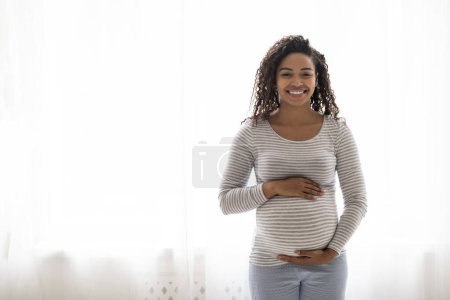 Foto de Esperando un bebé. Retrato de la señora embarazada negra feliz tocando tiernamente el vientre cerca de la ventana en casa, hermosa afroamericana esperando a la madre abrazando la panza y sonriendo, disfrutando del tiempo de embarazo - Imagen libre de derechos