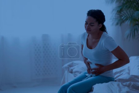 Photo for Unhappy young black woman suffering stomach pain in the night sick african american female sitting on bed at home, touching her belly, suffering from abdominal cramps, having stomachache, copy space - Royalty Free Image