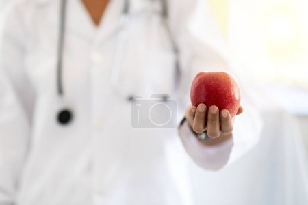 Photo for European middle aged lady doctor nutritionist in white coat recommends red apple in clinic interior, close up. Work, professional recommendation and health care, weight loss and diet - Royalty Free Image
