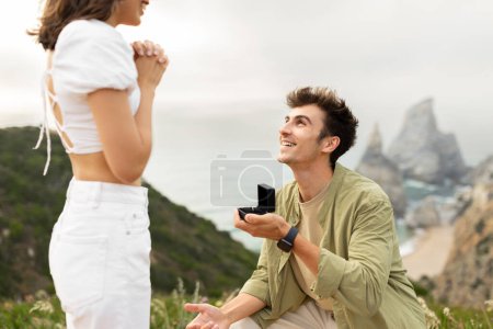 Photo for Happy young man standing on his knee and proposing to his girlfriend on a rocky cliff by the ocean shore, holding box with ring and looking at her beloved lady - Royalty Free Image