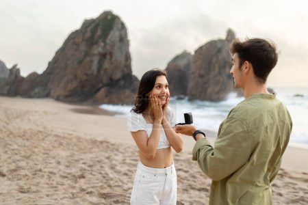 Photo for Marry me concept. Young guy making proposal to his surprised girlfriend with ring in gift box, standing on beach during romantic date on coastline, copy space - Royalty Free Image