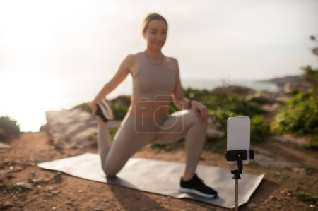 Photo for Happy millennial caucasian lady athlete in sportswear doing leg stretching exercise, shoots video on phone on sea beach. Sports blog outdoor, fitness app, body care and device, remote coach - Royalty Free Image