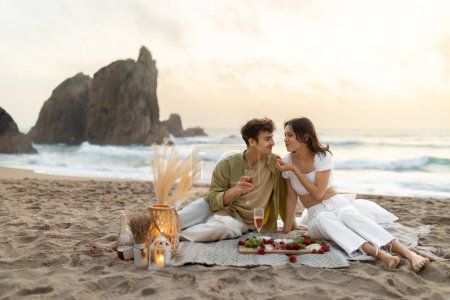 Foto de Pareja joven y romántica haciendo picnic en la playa de arena con vista al mar, bebiendo vino y comiendo frutas, disfrutando de una cita por la noche en la costa, espacio libre - Imagen libre de derechos