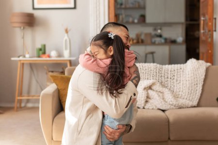 Téléchargez les photos : Petite fille japonaise embrassant son père exprimant amour et affection dans le salon moderne confortable à la maison. Le père embrasse sa petite princesse. Lien et connexion entre papa et enfant - en image libre de droit