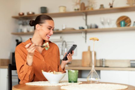Foto de Mujer europea feliz comiendo sabrosa ensalada y el uso de teléfono celular en la cocina mientras cenan, sentado en la mesa y mensajes de texto en el teléfono móvil, espacio para copiar - Imagen libre de derechos