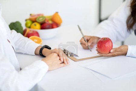 Photo for European middle aged lady doctor nutritionist in white coat write diet program to woman patient, recommends apple in clinic interior, cropped. Health care, treatment, weight loss with professional - Royalty Free Image