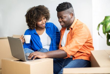 Photo for Happy Black Couple Using Computer After Moving To New Home, Smiling African American Spouses Sitting Among Cardboard Boxes In New Flat, Ordering Furniture Online, Purchasing Goods In Internet - Royalty Free Image