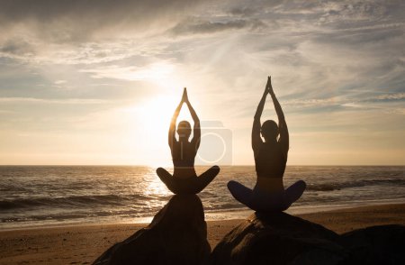 Foto de Hermanas gemelas caucásicas milenarias practican yoga en posición de loto, disfrutan de ejercicios de respiración en la playa de mar juntas. Entrenamiento matutino, deportes, atención médica y estilo de vida, espalda - Imagen libre de derechos