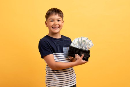 Photo for Pocket money, kids savings, financial literacy for children. Cheerful cute preteen kid handsome boy in casual outfit showing leather wallet full of cash dollar banknotes, smiling, yellow background - Royalty Free Image