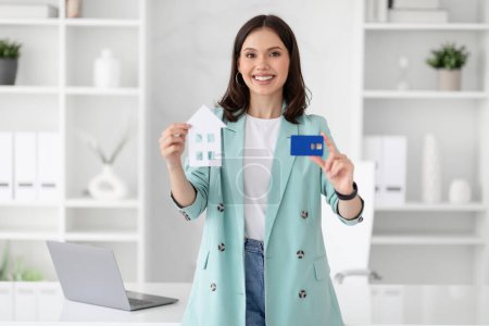 Photo for Cheerful young caucasian businesswoman in suit hold credit card and house in office interior. Investment recommendations, business, realtor work, profit, loan for real estate - Royalty Free Image