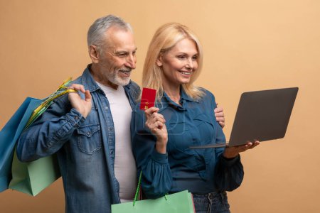 Photo for Joyful happy beautiful senior couple shopping online over beige background, elderly man and woman using computer laptop and bank credit cardm holding purchases. E-commerce, retail concept - Royalty Free Image